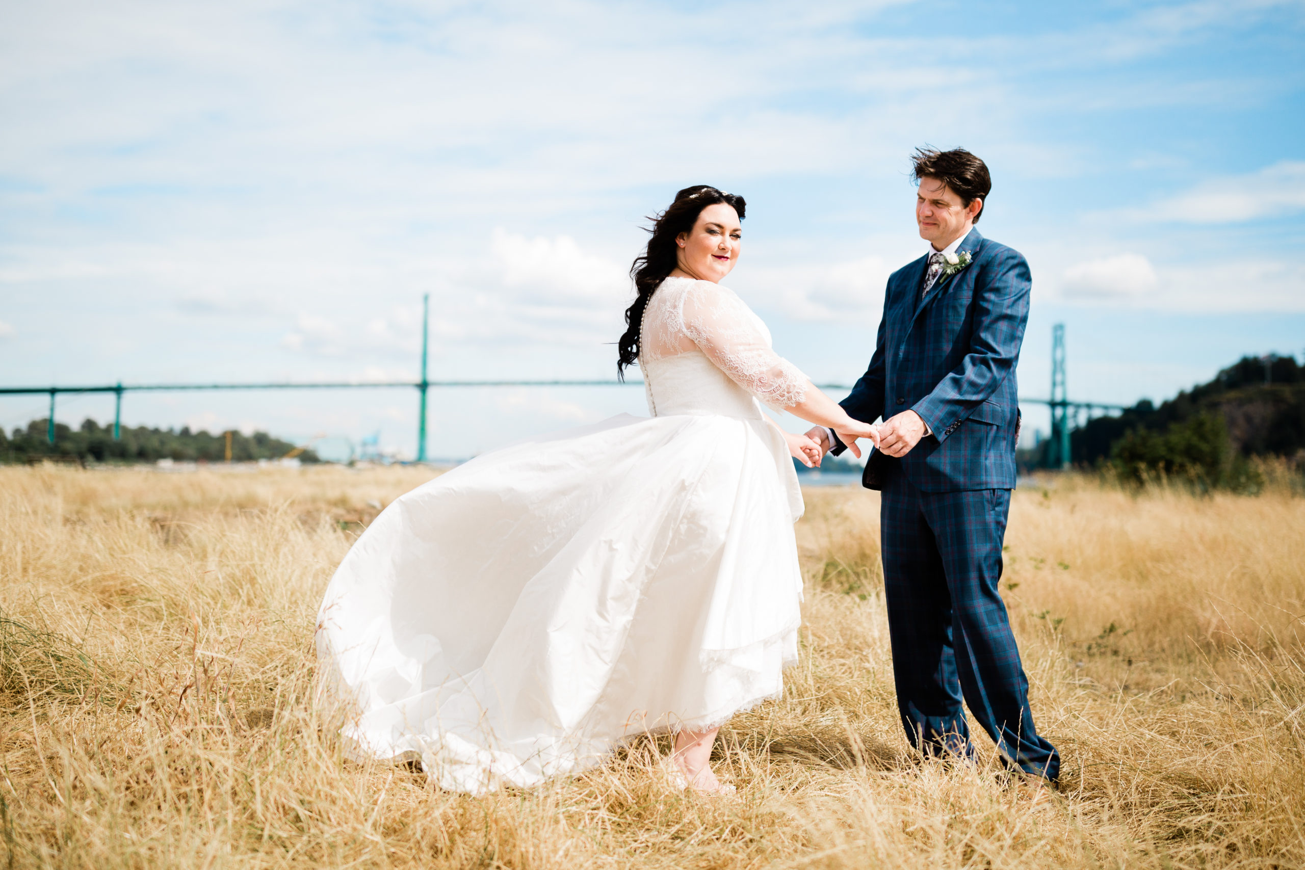 Bride and groom standing in field with Lions Gate bridge behind them.