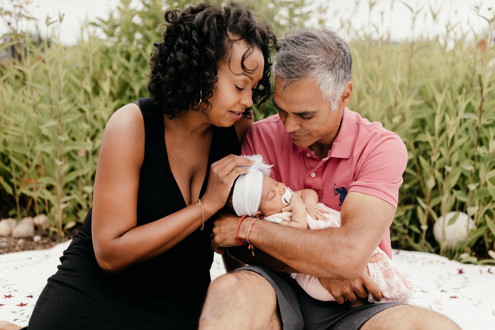 Mom and dad holding newborn daughter, sitting in the tall grass.